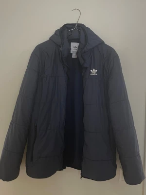 Mörkblå pufferjacka från Adidas - Säljer en mörkblå pufferjacka från Adidas med huva och dragkedja framtill. Jackan har Adidas-logga på bröstet och praktiska sidofickor med dragkedja. Perfekt för kalla dagar och har en clean, sportig vibe.