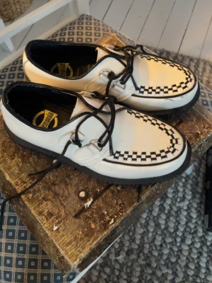 Demonia Creeper Storlek:40 - Säljer ett par Demonia Creeper-602 i vit och svart med schackrutigt mönster runt tån. Skorna har snörning med metallringar, platt sula och ovandel i skinn. Snygg kontrast mellan svart och vit, perfekt för dig som gillar alternativ stil.