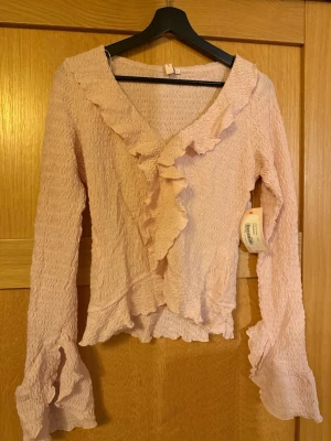 Ljusrosa omlottblus med volanger från Nelly - Säljer en ljusrosa omlottblus från Nelly med vågigt strukturmönster och volangdetaljer längs halsringningen och ärmsluten. Blusen har lång ärm och v-ringad hals. Perfekt för dig som gillar romantiska detaljer och mjuka färger.