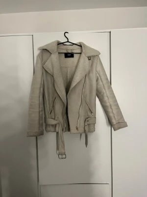 Beige teddyjacka med bälte från Bik Bok - Säljer en beige teddyjacka från Bik Bok med mjukt foder, bred krage och bälte i midjan. Jackan har dragkedja framtill, två snedställda fickor med dragkedja och detaljer i guld. Perfekt för kalla dagar och riktigt snygg till jeans.