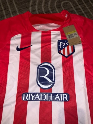 Rödvit randig Nike Atlético Madrid tröja - Säljer en officiell Atlético Madrid matchtröja från Nike med röda och vita ränder, klubbmärke på bröstet och sponsorloggor från Riyadh Air och Ria Money Transfer. Tröjan har korta ärmar och är tillverkad i polyester för en sportig känsla.