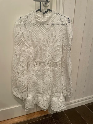Vit spetsklänning med volang - Supersöt vit kortklänning med broderad spets och volangdetaljer. Klänningen har långärmade puffärmar och lite högre hals med fina spetskanter. Perfekt studentklänning. Endast använd en gång.