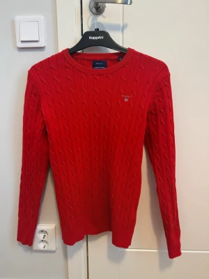 Röd kabelstickad tröja från GANT - Säljer en snygg röd kabelstickad tröja från GANT i stretchig bomull. Tröjan har rund hals, lång ärm och diskret broderad logga på bröstet. Perfekt för dig som gillar klassisk stil med en modern twist. Passar dig som vill ha en normal passform.