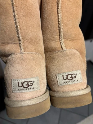 Beige UGG boots - Säljer ett par klassiska UGG boots i beige mocka med mjukt fårskinnsfoder. Skorna har rund tå, platt sula och tydliga sömmar längs skaftet. Perfekta för kalla dagar och riktigt sköna att dra på sig. UGG-logga bak på hälen och mönstrad yttersula.