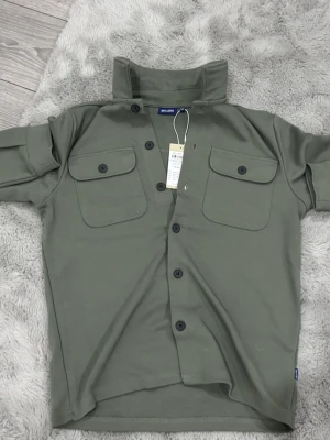 Grön overshirt från Only & Sons - Snygg grön overshirt från Only & Sons med klassisk krage och två stora bröstfickor med knappar. Jackan har knäppning hela vägen och är tillverkad i mjukt bomullsmaterial. Perfekt att slänga på över en t-shirt för en chill look. Helt oanvänd och pris kan diskuteras