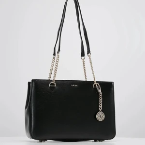 Svart axelväska från DKNY - Svart axelväska från DKNY med kedjedetaljer i silver och ett hängande DKNY-emblem. Rektangulär form och är tillverkad i skinn. 