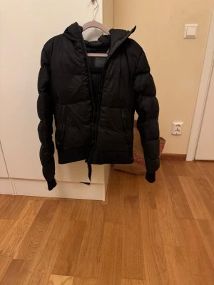 Svart pufferjacka med huva, strl 38 - Svart pufferjacka i storlek 38 med huva och dragkedja framtill. Jackan har två fickor med dragkedja och ribbade muddar vid ärmslut. Perfekt för kalla dagar och har en snygg, lite puffig look. Och en röd hoodie i storlek S