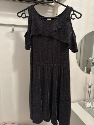 Svart glittrig kortklänning med volang - Supersnygg svart kortklänning med glittrigt tyg och volangdetalj över axlarna. Klänningen har öppna axlar och en mjuk, flowy kjol. Perfekt för dig som gillar att sticka ut med lite extra glitter.