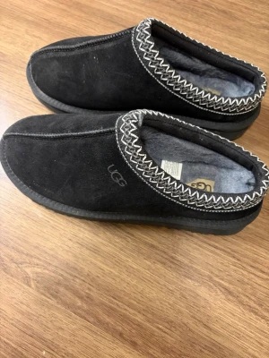 Svarta UGG slip-in skor med ullfoder - Mysiga slip-in skor från UGG i svart mocka med fluffigt grått ullfoder. Snyggt mönstrad kant i vitt och svart runt öppningen. Platt sula med UGG-logga undertill. Perfekta för chill dagar hemma eller när du vill ha det extra bekvämt.