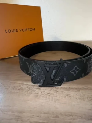 L och v bälte -  skärp med klassiskt monogrammönster i svart och grått. Snygg logga i matt svart metall på spännet. Skärpet är tillverkat i äkta skinn med brunt insida och detaljerad söm. Perfekt accessoar för att lyfta din outfit.