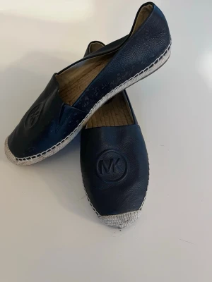 Svarta loafers från Michael Kors - Snygga svarta loafers från Michael Kors med MK-logga på ovansidan. Skorna har en rund tå, platt sula och är tillverkade i skinn med flätad detalj vid tån. Insidan har en beige, repmönstrad sula som ger en chill vibe. Perfekt för dig som gillar stilrena och klassiska skor.