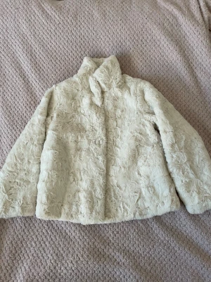 Krämvit pälsjacka för barn från H&M  - Säljer en fluffig och mysig krämvit pälsjacka för barn från H&M med hög krage och långa ärmar. Jackan har ett glansigt satin-foder och stängs med knappar framtill. Perfekt för dig som vill ha en varm och stylish look under hösten och vintern.