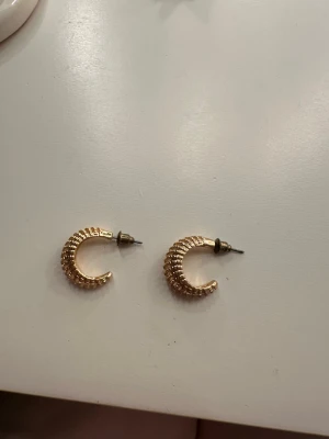 Guldiga ribbade örhängen - Säljer ett par chunky guldiga örhängen med ribbad yta och halvcirkelformad design. De är i metall och har ett coolt, texturerat mönster som ger extra edge till din look. Perfekta om du gillar statement-smycken och vill sticka ut lite extra.