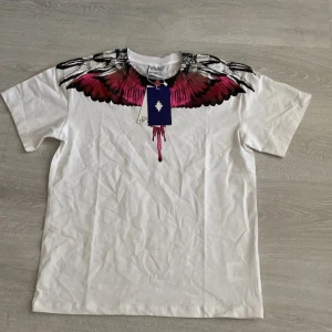 Marcelo Burlon tshirt - (Passar även folk som har storlek S) Snygg vit ärmlös t-shirt från med ikoniskt tryck av rosa och svarta vingar över axlar och bröst. T-shirten har rund halsringning och är tillverkad i mjuk bomull. Perfekt för dig som vill sticka ut med en cool och unik design.