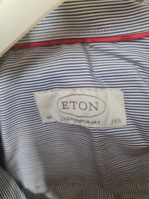 Randig skjorta från Eton, blå/vit - Snygg skjorta från Eton i blått och vitt med tunna ränder. Klassisk krage, vita knappar och diskret röd detalj på insidan av kragen. Skjortan är långärmad och har en modern passform som funkar till många stilar. Perfekt för dig som gillar stilrena plagg med lite edge.