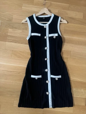 Svart och vit kort klänning från SHEIN - Snygg svart ärmlös kortklänning från SHEIN med vita detaljer och dekorativa silverfärgade knappar framtill. Klänningen har rund halsringning och två fickor med vita kanter. Materialet är ribbat och ger en cool struktur.