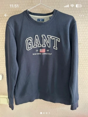  sweatshirt från GANT - Säljer en mörkblå sweatshirt från GANT med klassisk logotyp och amerikansk flagga broderad framtill. Tröjan har rund hals, långa ärmar och är tillverkad i mjuk bomull med lite polyester för extra komfort. Perfekt för dig som gillar stilren och sportig look.