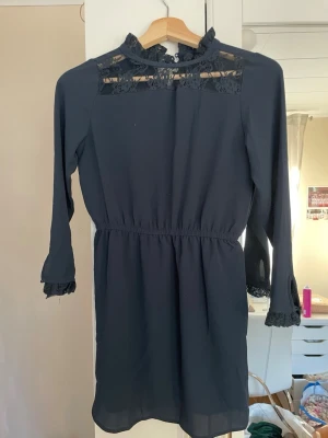 Mörkblå spetsklänning för barn från Name It - Snygg mörkblå kortklänning för barn från Name It med långärmade ärmar och spetsdetaljer vid halsringning och ärmslut. Klänningen har elastisk midja och en liten volang vid kragen. Perfekt för dig som gillar stilrena detaljer och spets.