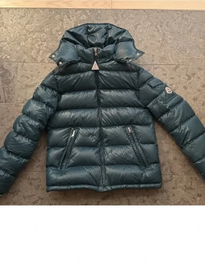Mörkgrön jacka från Moncler - Säljer en mörkgrön pufferjacka från Moncler med huva och två dragkedjeförsedda fickor. Jackan har quiltad design och Moncler-logga på ärmen. Materialet är glansigt och känns riktigt lyxigt, perfekt för kalla dagar. tveka inte på o höra av er!