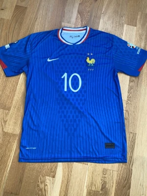Blå Frankrike Mbappé fotbollströja - Säljer en blå Frankrike fotbollströja från Nike med nummer 10 och Mbappé på ryggen. Tröjan har vita och röda detaljer vid kragen, FFF-logga med tupp på bröstet och officiella EM-märken på ärmarna. Tillverkad i lätt polyester med mönstrad design och rund hals.