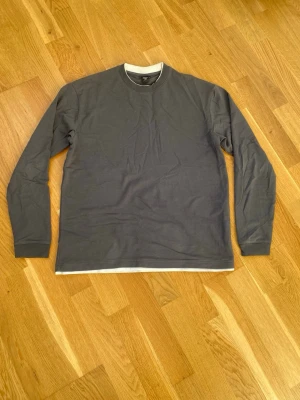 Mörkgrå långärmad topp - En enkel och stilren mörkgrå långärmad topp med rund halsringning. Tillverkad i mjuk bomull och har en klassisk passform. Perfekt att matcha med jeans eller kjol för en clean look.