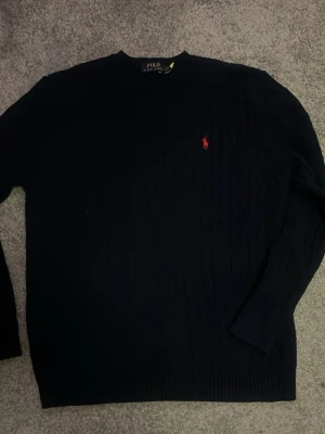 Mörkblå stickad tröja från Polo Ralph Lauren - Snygg mörkblå stickad tröja från Polo Ralph Lauren med klassisk kabelstickning och röd broderad logga på bröstet. Tröjan har rund hals och långa ärmar, perfekt för att hålla värmen under kyliga dagar.