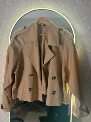Beige kort trenchcoat med knappar - Säljer en beige kort trenchcoat från H&M med dubbelknäppning och breda ärmar. Perfekt vår / höst jacka! 