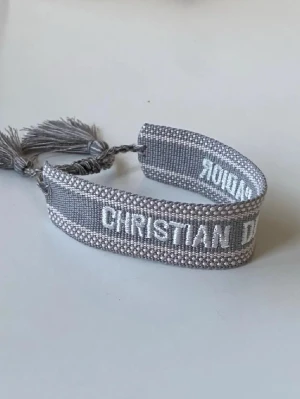 Grå armband från Christian Dior - Säljer ett snyggt grått armband från Christian Dior med broderad logga i vitt. Armbandet är gjort i ett stickat material och har justerbar snörning med tofsar i ändarna. Perfekt accessoar för att lyfta din outfit.
