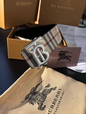 Burberry vändbart bälte med giftbox set - Säljer en Burberry väst med klassiskt rutig mönster i beige, svart och röd. Västen har ett stort metallspänne med Burberrys B-logga framtill. Materialet är exklusivt skinn och designen är tidlös med lyxig känsla. Perfekt för dig som vill sticka ut med en ikonisk detalj.