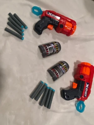 Röd X-Shot väst med patronhållare - Cool väst från X-Shot i rött med svarta och orange detaljer. Västen har integrerade patronhållare och medföljande patroner i grått med blå toppar. Perfekt för att bära och förvara skumdart-pistoler och extra ammunition under lek. Västen har en sportig design och är tillverkad i syntetmaterial.