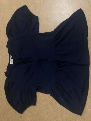 Mörkblå blus med knyt från Gina Tricot - Säljer en mörkblå blus från Gina Tricot i mjuk bomull. Blusen har korta puffärmar och två knytar framtill. Den har en volang nedtill. Perfekt att styla med jeans eller kjol. I storlek 164, aldrig använt.