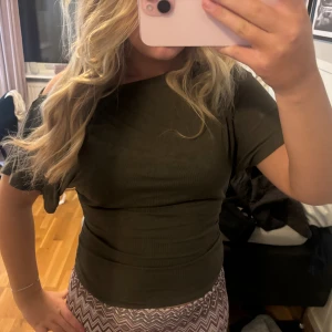 Mörkgrön offshoulder topp - Trendig mörkgrön topp med off sopulder-design och tight passform. Toppen är ärmlös på ena sidan och har kort ärm på andra, vilket ger en asymmetrisk look. Perfekt att styla med höga jeans eller kjol för en cool vibe.