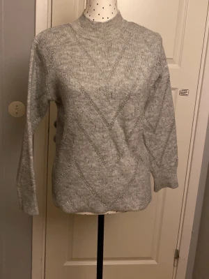 Ljusgrå stickad tröja med glitter - Mysig ljusgrå stickad tröja med diskreta silvriga glittertrådar i ett sicksackmönster. Tröjan har hög hals, långa ärmar och ribbade muddar. Perfekt för dig som vill ha något både bekvämt och snyggt till hösten eller vintern. Lite glittertrådar som sticker ut. 
