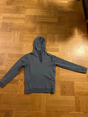 Blå hoodie från Lyle & Scott - Snygg blå hoodie från Lyle & Scott med klassisk gul logga på bröstet. Hoodien har huva med snören och en stor magficka framtill. Tillverkad i mjukt material som passar perfekt för en chill och avslappnad stil.