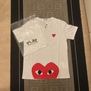 Vit Comme des Garçons Play t-shirt S - Storlek M men passformen är S. Snygg vit t-shirt från Comme des Garçons Play med ikoniskt rött hjärta med ögon på bröstet och ett stort hjärta med ögon längst ner. Klassisk rund hals och korta ärmar. Materialet är mjuk bomull och passformen är normal. Perfekt statement-plagg för dig som gillar streetstyle.