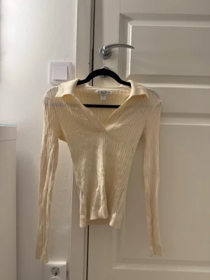 Ribbad beige långärmad topp  - Trendig ribbad topp från AMIEU i beige med lång ärm och snygg krage, sitter tight. V-ringad framtill och figurnära passform som ger en clean och stilren look. Perfekt att matcha med jeans eller kjol för en enkel men chic outfit. 