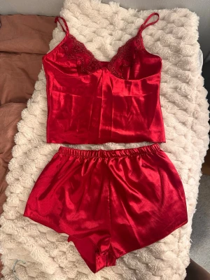 Rött satinsätt med spetsdetaljer - Snyggt rött satinsätt med linne och shorts. Linnet har tunna axelband och spetsdetaljer vid bysten för en extra feminin touch. Shortsen har resår i midjan och matchar linnet perfekt. Materialet är glansigt och mjukt, vilket ger en lyxig känsla. Litet hål som säkert går att laga, har bara inte orkat testa!
