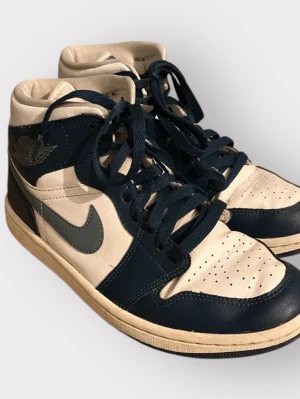 Nike Air Jordan 1 Mid blå/vit - Snygga Nike Air Jordan 1 Mid sneakers i marinblått och vitt skinn. Klassisk siluett med rund tå, snörning och ikoniska Jordan-loggan på sidan. Perfekta för dig som vill ha en stilren och sportig look.