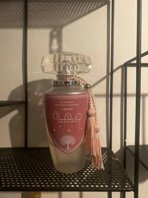 Lattafa Mohra Eau De Parfum - Lattafa Mohra Eau De Parfum i en elegant glasflaska med frostad finish och en stor, kristalliknande kork. Flaskan har en rosa etikett med vita detaljer och en dekorativ rosa tofs som hänger på sidan. Perfekt för dig som gillar lyxiga och unika dofter.