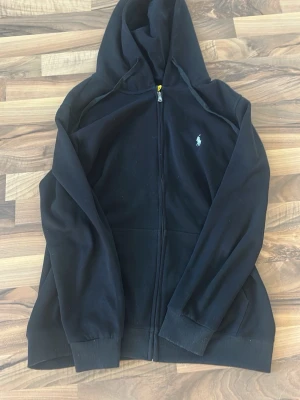 Svart hoodie från Ralph Lauren - Klassisk svart hoodie med dragkedja från Ralph Lauren. Har huva med dragsko, ribbade muddar och fickor framtill. Liten broderad logga i vitt på bröstet. Perfekt för en chill och stilren look. Storlek Xl men passar den som har L bra 