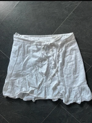 Vit omlottkjol med volang och knyt - Söt vit kort omlottkjol med volangkant och knytband i midjan. 