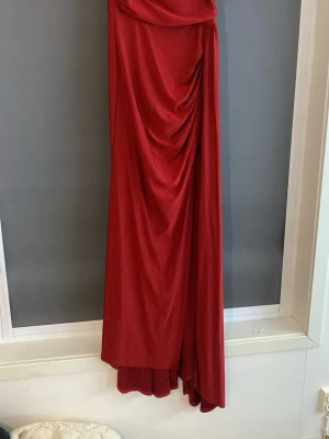 Röd långklänning med drapering - Elegant röd långklänning med vacker drapering framtill. Klänningen har en figurnära passform och ett mjukt, följsamt tyg som ger ett snyggt fall. Perfekt för speciella tillfällen när du vill sticka ut.