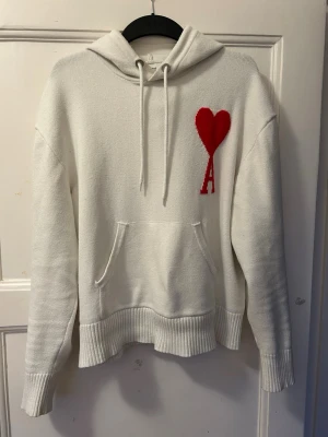 Vit hoodie från Ami Paris med rött hjärta - Säljer en vit hoodie från Ami Paris med ett stort rött hjärta och bokstaven A broderat på bröstet. Hoodien har huva med dragsko, magficka och ribbade muddar vid ärmar och nederkant. Tillverkad i mjukt stickat material som känns lyxigt och skönt. Liten fläck på midgan.