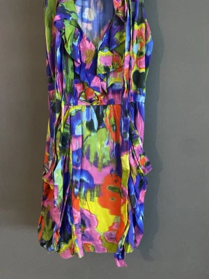 Färgstark kortklänning med volang - Unik kortklänning med färgstarkt abstrakt mönster i lila, blått, grönt, gult och rött. Klänningen har volangdetaljer vid ringningen och en lätt omlottkänsla framtill. Perfekt för dig som vill sticka ut med en lekfull och kreativ stil.