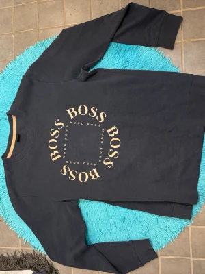 Svart sweatshirt från Hugo Boss - Svart sweatshirt från Hugo Boss med rund hals och stort BOSS-tryck i beige på bröstet. Tröjan har långa ärmar och ribbade muddar vid ärmslut och nederkant. Perfekt för en avslappnad och stilren look.