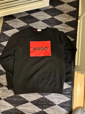 Svart sweatshirt från Hugo - Svart sweatshirt från Hugo med en stor röd logga på bröstet. Tröjan har rund halsringning, långa ärmar och ribbade muddar vid ärmslut och nederkant. Tillverkad i mjukt bomullsmaterial för en skön och avslappnad look.