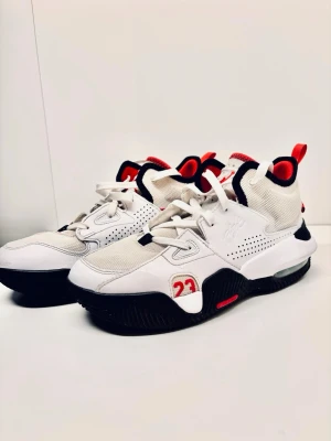 Vita Jordan sneakers med röda detaljer - Snygga vita Nike Jordan sneakers med röda och svarta detaljer. Skorna har snörning, meshpaneler och nummer 23 broderat på sidan. Mellanhög modell med vadderad häl och robust sula för extra komfort. Perfekta för dig som gillar streetwear och basketstil.