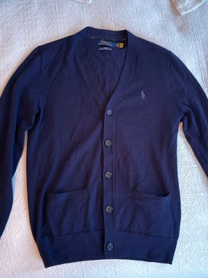 Mörkblå cardigan från Polo Ralph Lauren - Klassisk mörkblå cardigan från Polo Ralph Lauren med V-ringning, knäppning framtill och två fickor. Tröjan har diskret logga broderad på bröstet och är tillverkad i mjukt material. Perfekt för lager på lager och stilren look.