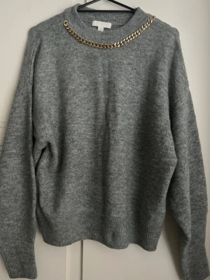 Grå stickad tröja med guldkedja H&M - Snygg grå stickad tröja från H&M i storlek XS. Tröjan har en unik detalj med en guldfärgad kedja runt halsringningen, ribbade muddar och avslappnad passform. Perfekt för dig som vill ha något extra till din vardagsstil. Passar både till jeans och kjol.  Storlek: XS Färg: Grå Märke: H&M Skick: Mycket bra – använd få gånger, inga skador eller noppor.