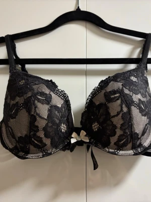 Svart spets-bh med rosett - Snygg svart bh med blommig spets och vadderade kupor. Dekorerad med en liten beige rosett framtill och diskret mesh under spetsen. Axelbanden är justerbara för bästa passform. Perfekt för dig som gillar en feminin och elegant stil.